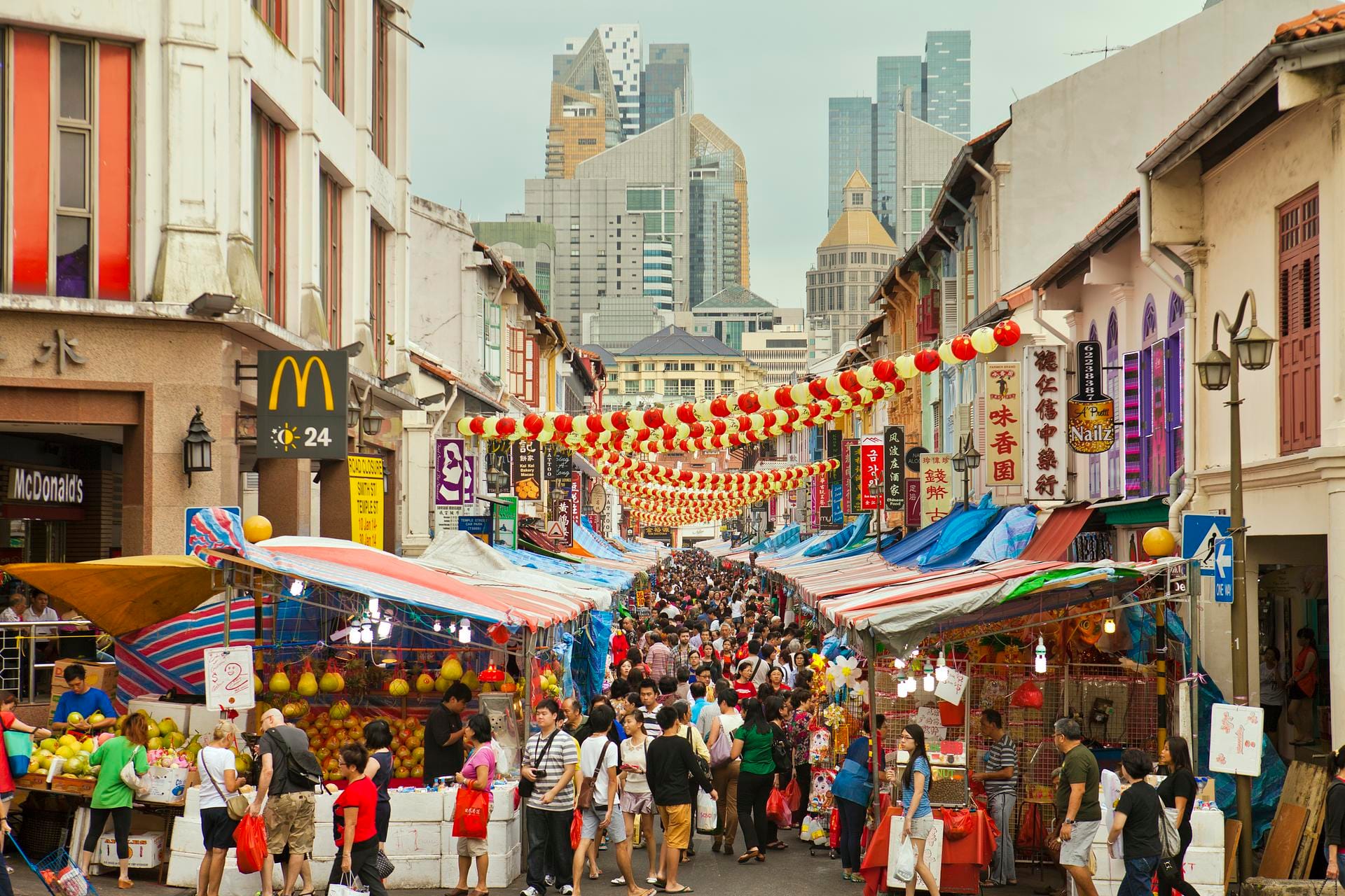 Shophouses and lanterns line a busy street in Chinatown, with skyscrapers in the background